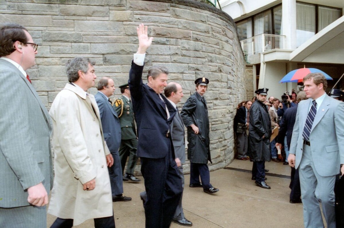 Ronald Reagan máva tesne predtým, ako ho strelia. Zľava sú Jerry Parr v bielom kabáte, ktorý natlačil Reagana do limuzíny, tlačový tajomník Bieleho domu James Brady, ktorý bol vážne zranený výstrelom do hlavy.