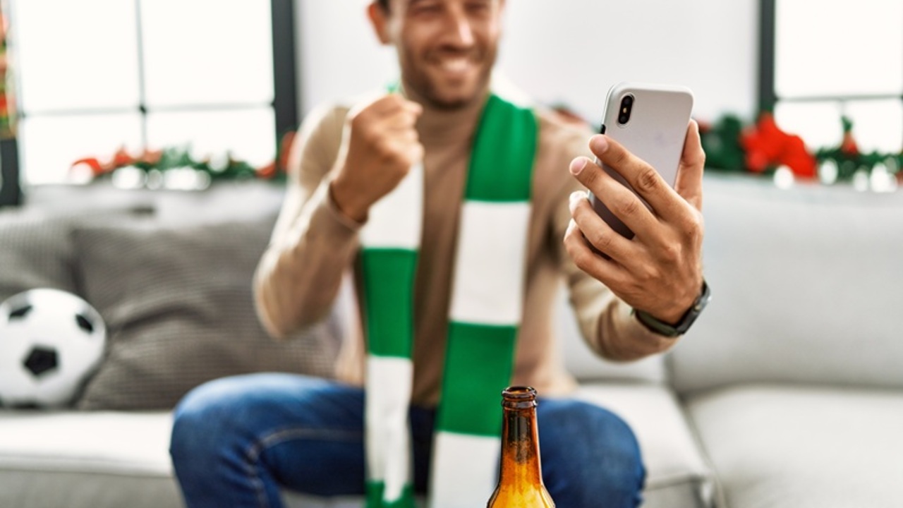 young-hispanic-man-supporting-soccer-match-using-smartphone-sitting-by-christmas-decor-home.jpg